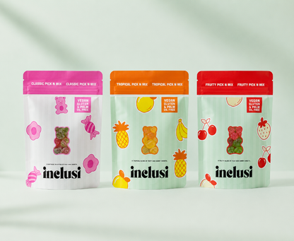 Three stand-up pouches of Inelus gummy bear candy mixes lined up on a light gray background with soft shadows. The left pouch is white with a pink top and pink fruit illustrations, the middle pouch is white with an orange top and tropical fruit illustrations, and the right pouch is white with a red top and strawberry illustrations.
