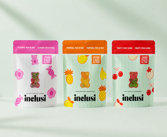 Three stand-up pouches of Inelus gummy bear candy mixes lined up on a light gray background with soft shadows. The left pouch is white with a pink top and pink fruit illustrations, the middle pouch is white with an orange top and tropical fruit illustrations, and the right pouch is white with a red top and strawberry illustrations.